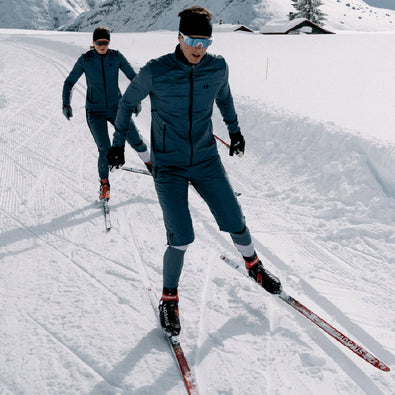 Two skiiers on the ski tracks, wearing Trimtex Primaloft Jacket and Shorts over the Ace Racesuit.